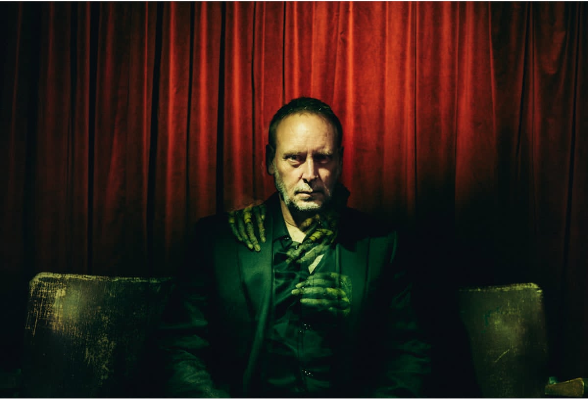 Kevin Kopfstein seated before a crimson curtain as spectral green hands rest upon his shoulders.