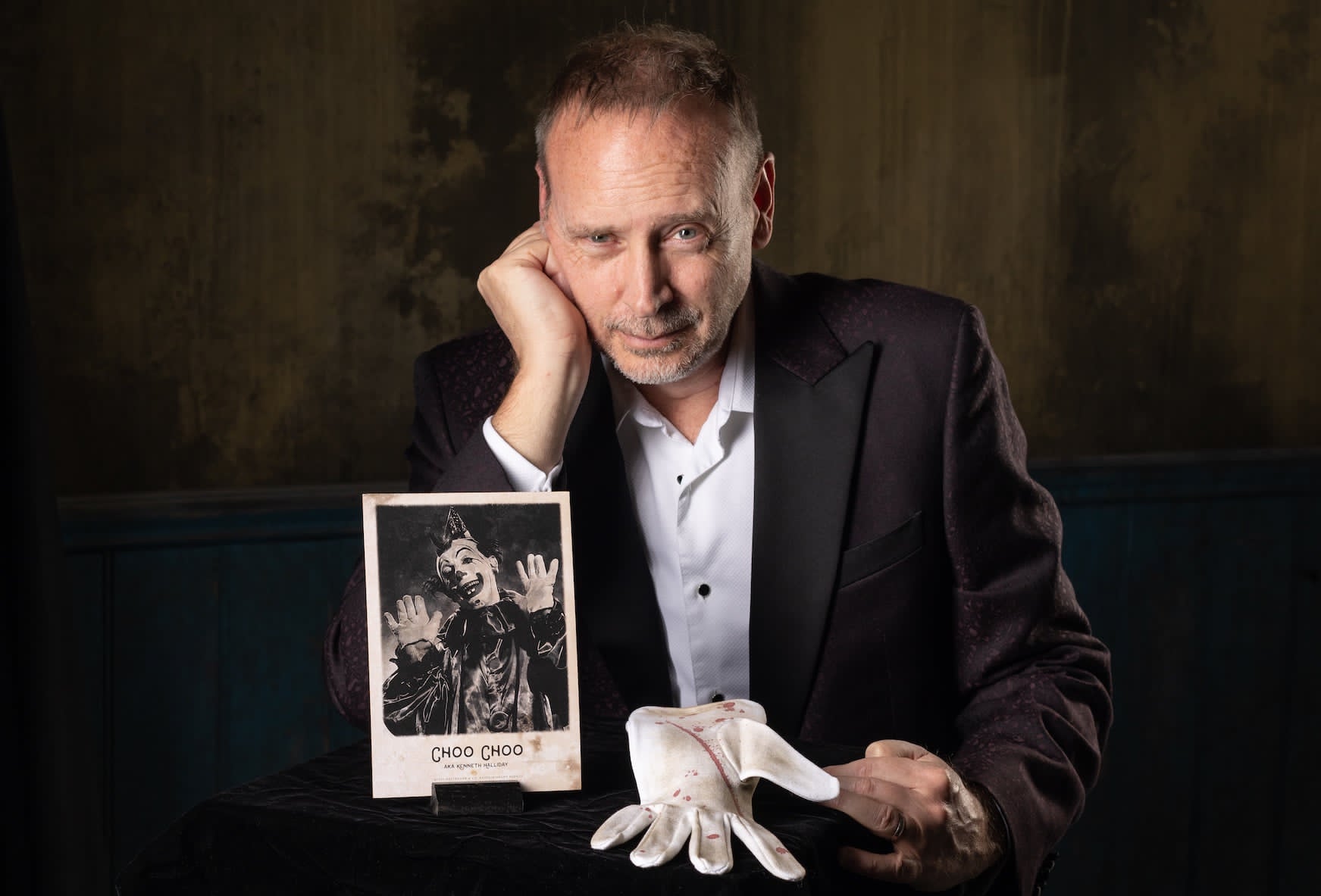 Kevin Kopfstein resting his head on his hand beside an antique haunted clown photograph and a blood-spotted glove.