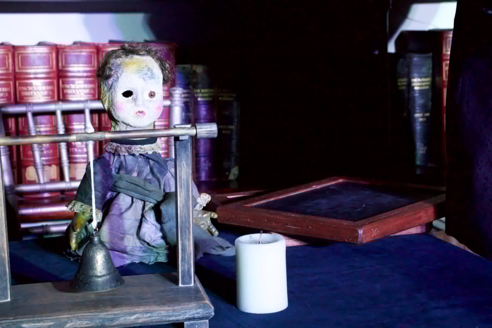 An antique haunted doll with hollow eyes restrained on a wooden stand beside a candle and old leather books.