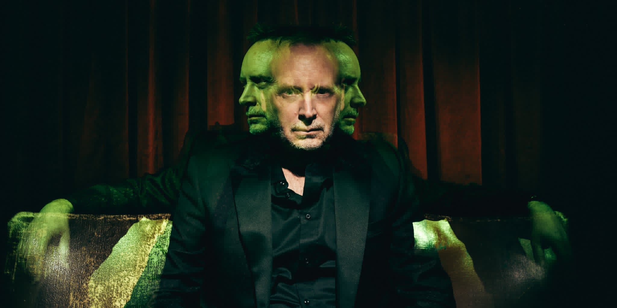 Kevin Kopfstein lit in eerie green and crimson light against a dark velvet curtain, his face multiplied as if surrounded by spirits.